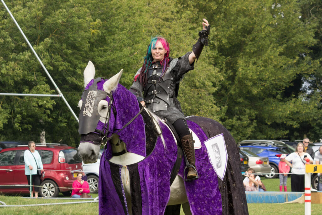 A person with pink and blue hair, wearing medieval armour, waves a heartfelt farewell whilst sitting on a white horse draped in purple and black fabric at an outdoor event with spectators and parked cars in the background.