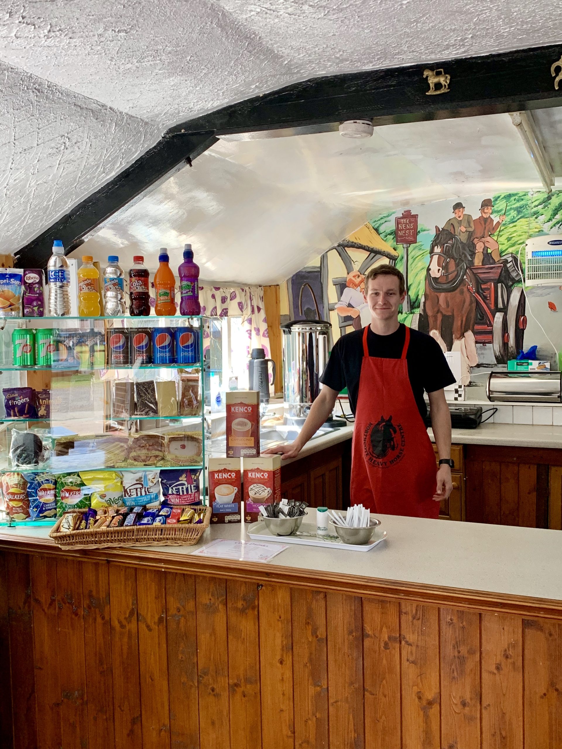 A smiling person wearing a red apron stands behind the counter of a small café, with snacks, drinks, and coffee supplies displayed. A mural with horses and people is painted on the wall behind them.