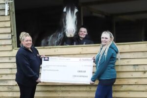 Three women smile while holding a large cheque for £800 in front of a wooden stable, with a horse and one woman inside the stable window, and the other two women standing outside.