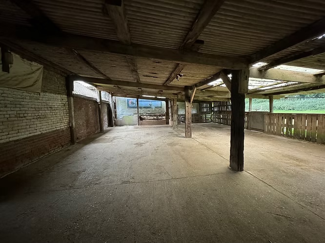 An empty, open-sided barn with a concrete floor, exposed wooden beams, brick walls, and sunlight streaming through clear roof panels and side slats. The barn opens to greenery outside.