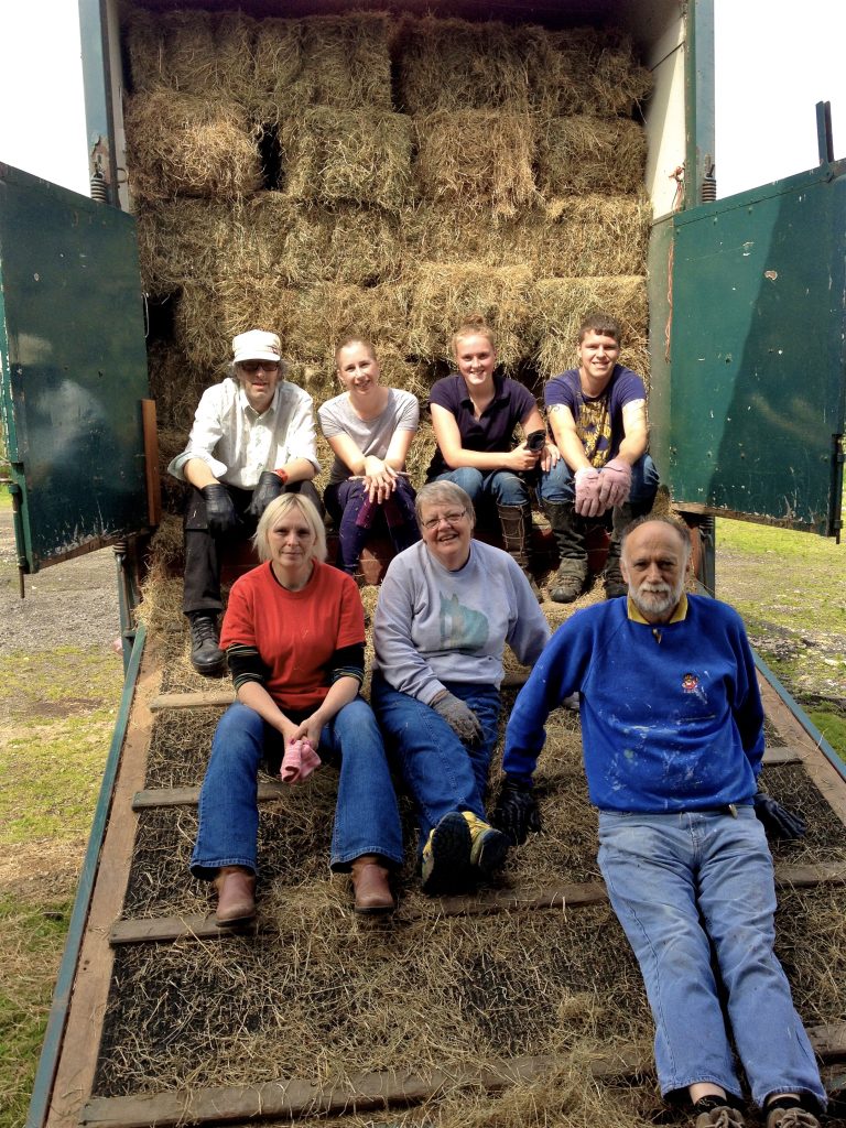 Seven people sit and pose in front of an open trailer filled with stacked hay bales. Four are seated on the edge of the trailer, and three sit on the ramp, all smiling outdoors on a sunny day.