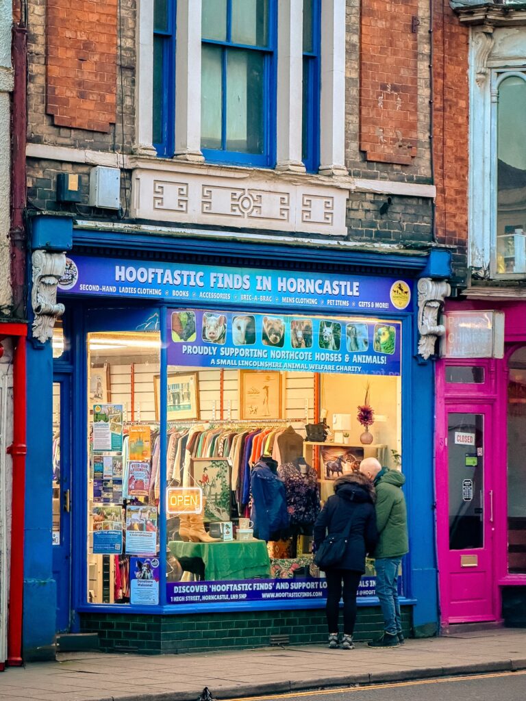 Two people stand outside Hooftastic Finds in Horncastle, a second-hand shop supporting horses and animals. The blue shopfront’s window showcases clothes, accessories, and colourful treasures awaiting discovery.