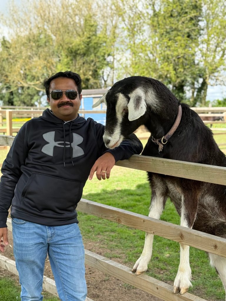 A man in a black hoodie and sunglasses stands by a wooden fence, smiling, while a goat with a collar leans over the fence and nuzzles his arm. Trees and green grass are visible in the background.