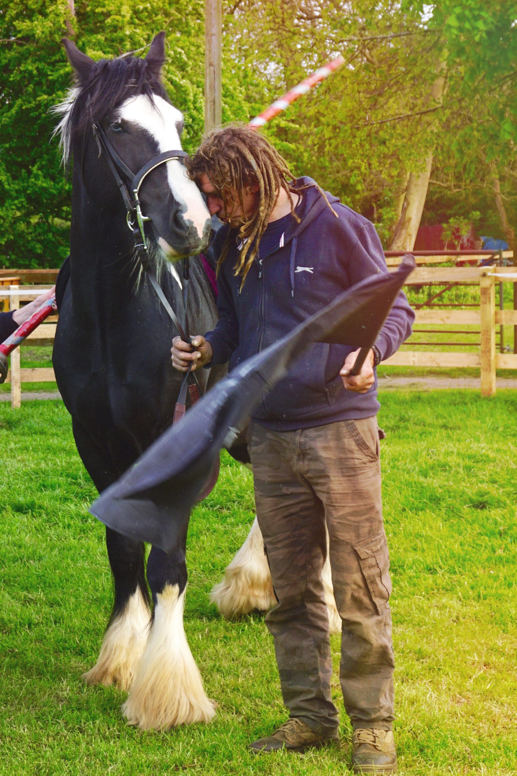 A person with dreadlocks stands beside a large black and white horse on grass, holding the reins and a black cloth, with wooden fences and green trees in the background.