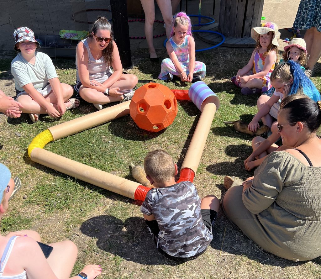 A group of children and adults sit in a circle on grass, surrounding a triangular structure made of tubes and a large orange ball in the centre, enjoying an outdoor activity on a sunny day.