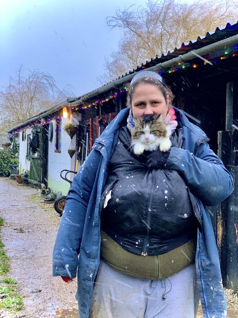 A person in a blue jacket holds a fluffy cat near their face, standing outside by a decorated building with colourful fairy lights. Snow is lightly falling, and the ground is wet and muddy.