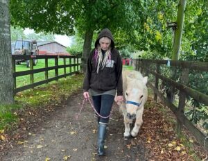 A woman walks a small horse near the stables, their quiet bond offering comfort after a devastating loss.