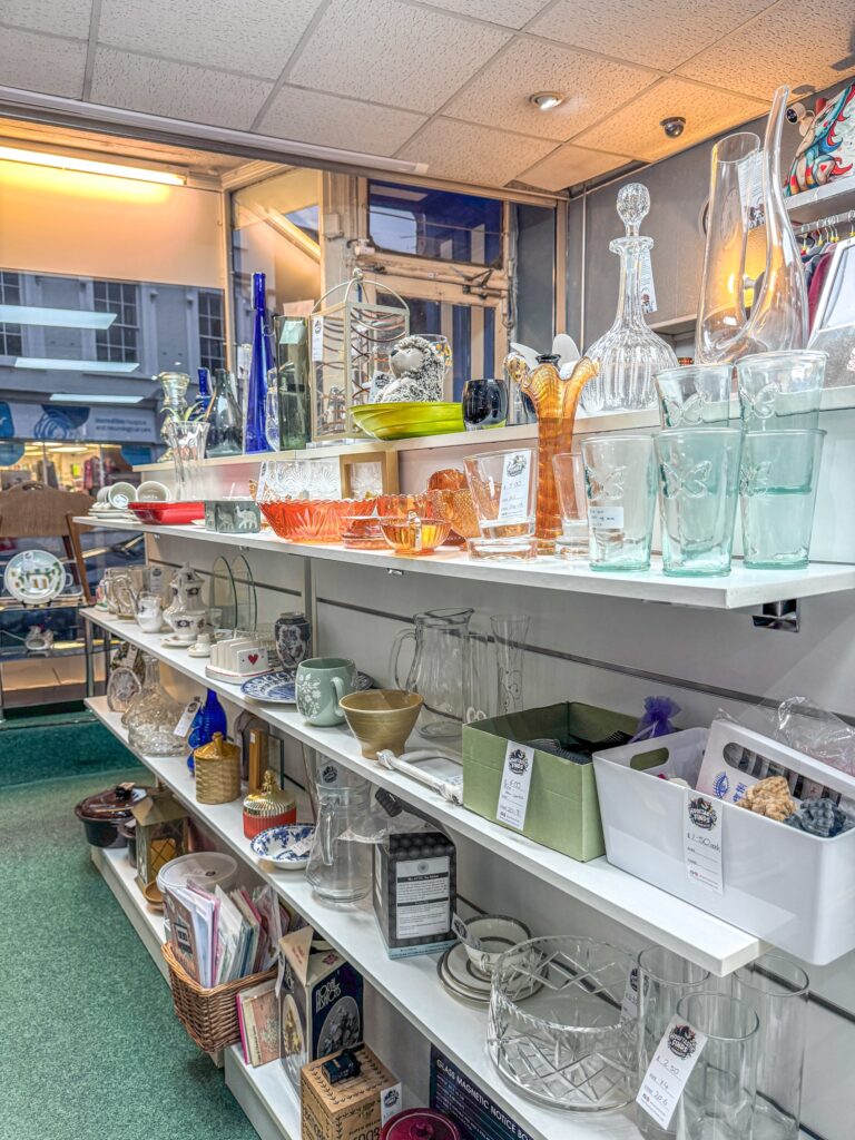 Shelves in Hooftastic Finds charity shop display various glassware, crockery, vases, cups, and decor items. In the background, more shelves and treasures await discovery, with a window reflecting the outside street scene.